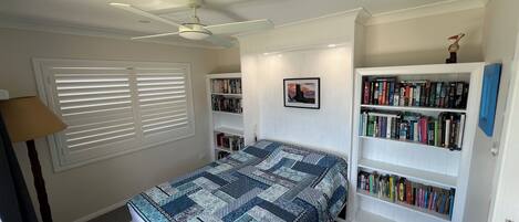 Een slaapkamer met een bed, een plafondventilator, een boekenplank en een raam met jaloezieën.