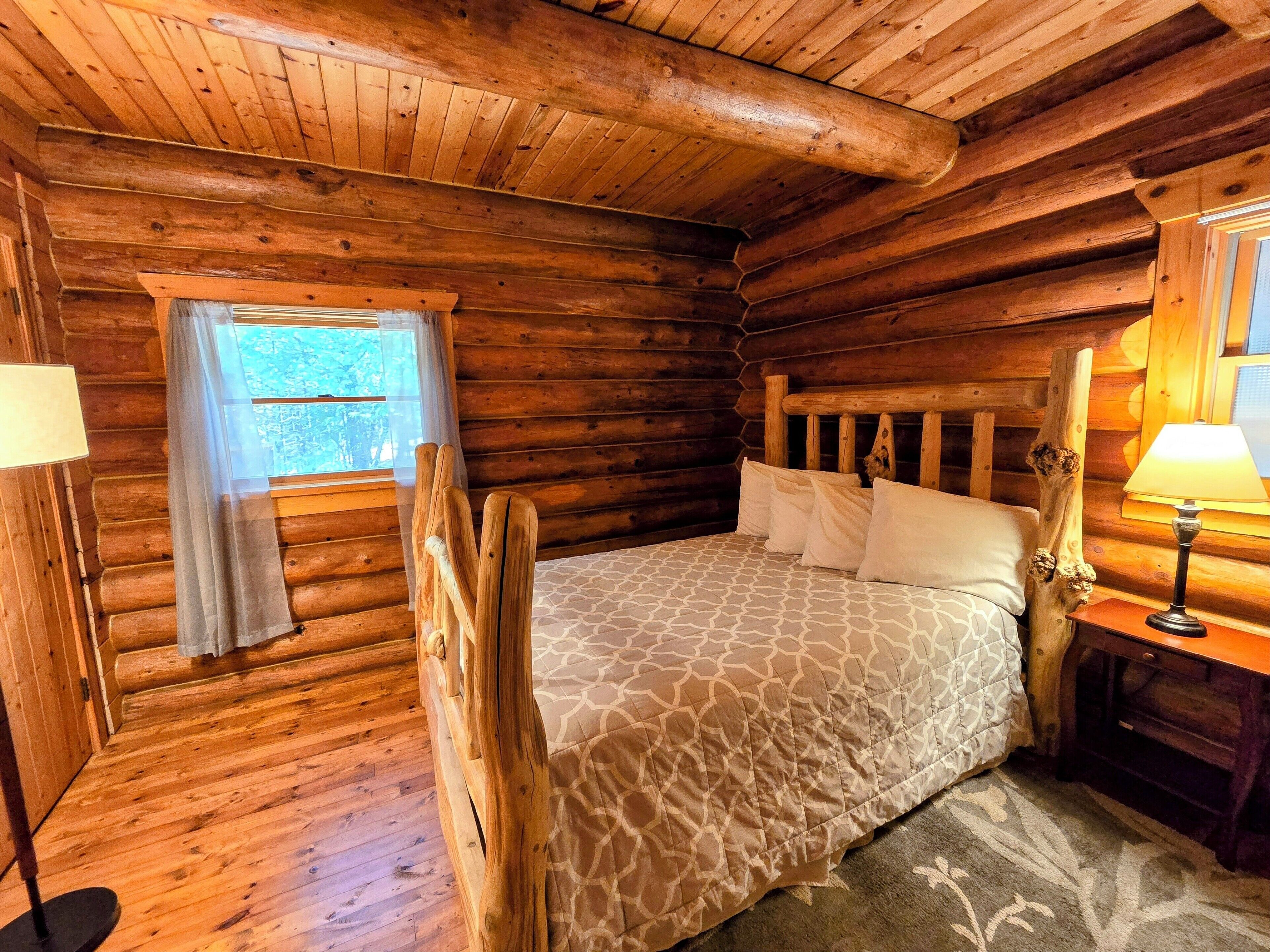 Camera da letto in una baita di legno con un letto, un comodino, una lampada, una finestra con tende e un pavimento in legno.