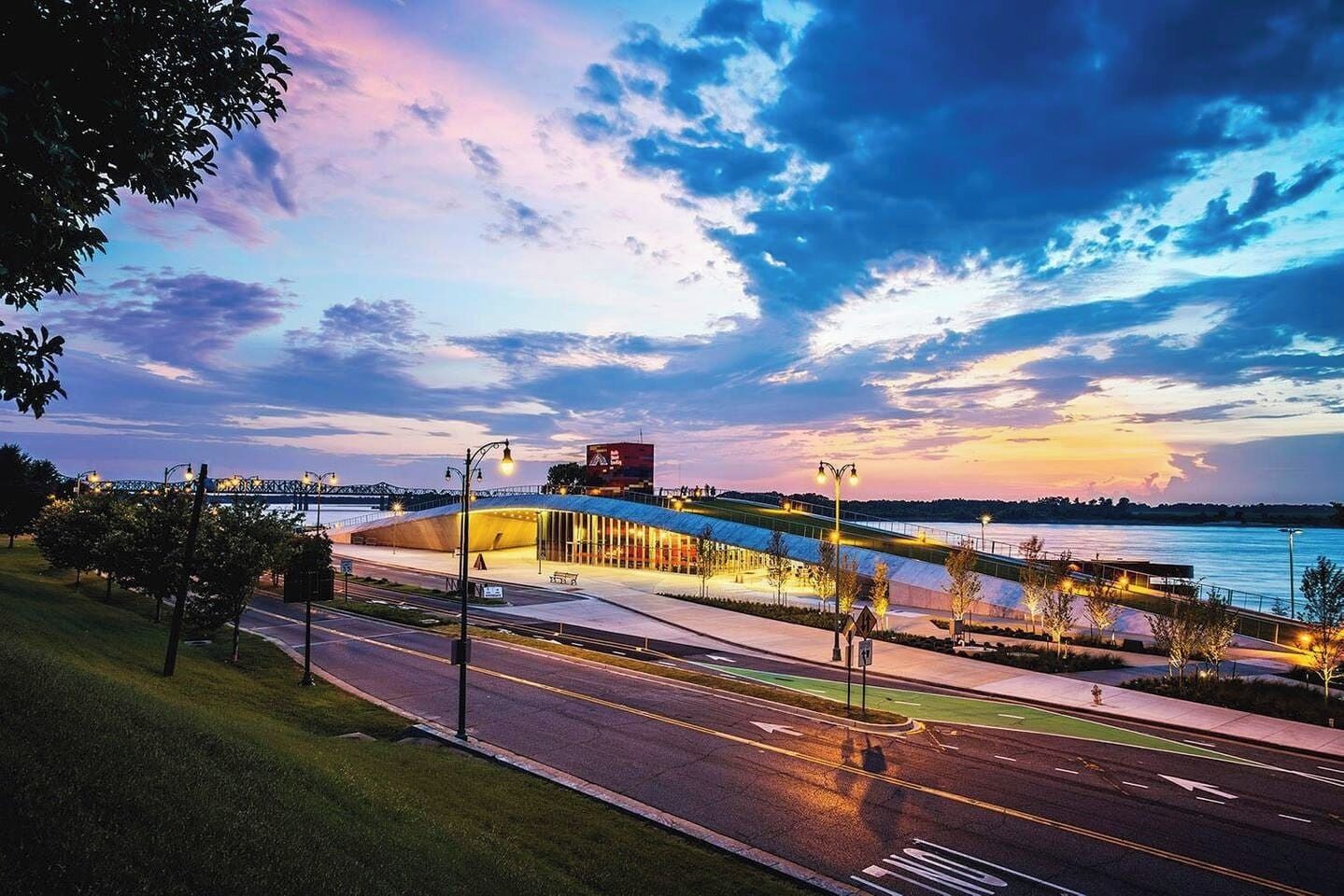 Un edificio moderno con techo curvo, un paseo peatonal y vista a una masa de agua al atardecer.