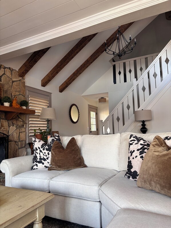 A living room with a sectional sofa, a wooden coffee table, a stone wall, a staircase with a white railing, and a chandelier.