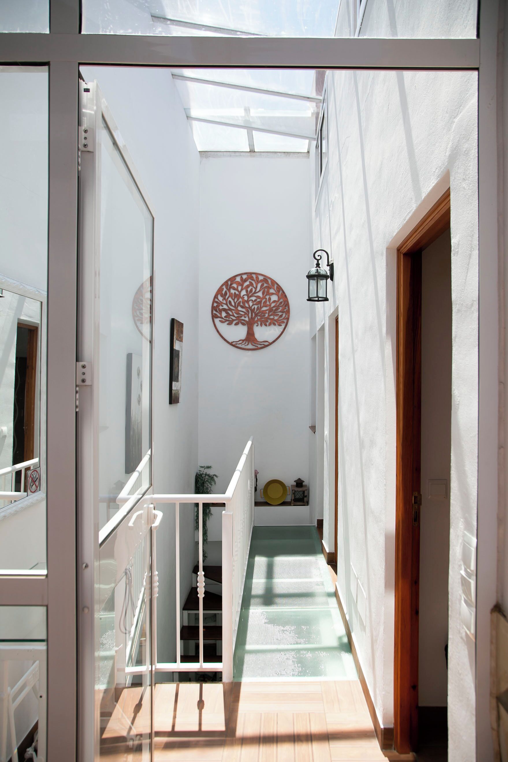 Un pasillo estrecho con suelo de madera, paredes blancas y un portall vidrio que da a un balcón. Se observa una decoración mural con forma de árbol y un farol.