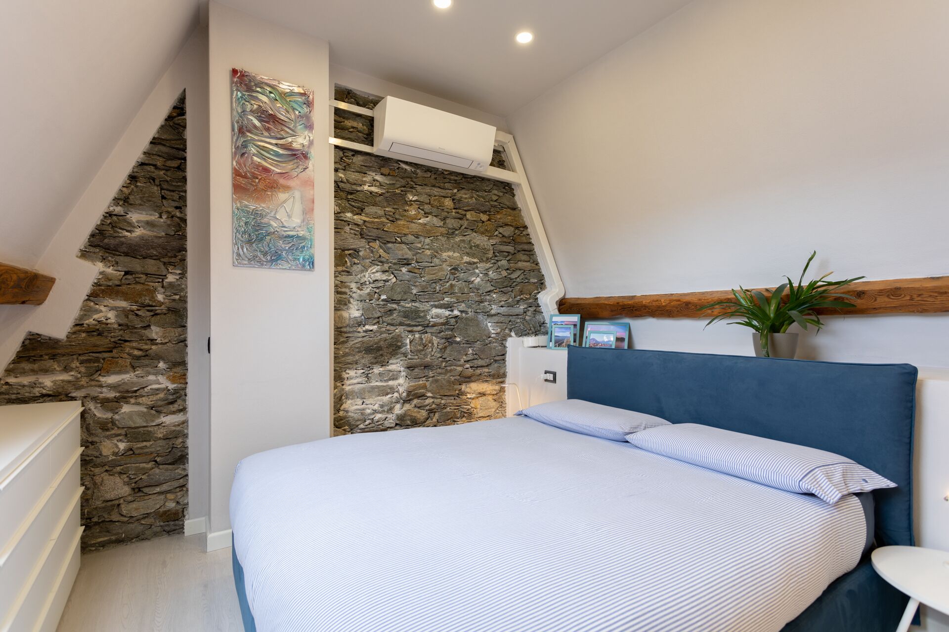 Un dormitorio con pared de piedra, cama con ropa de cama a rayas, mesita de noche y una planta en maceta.