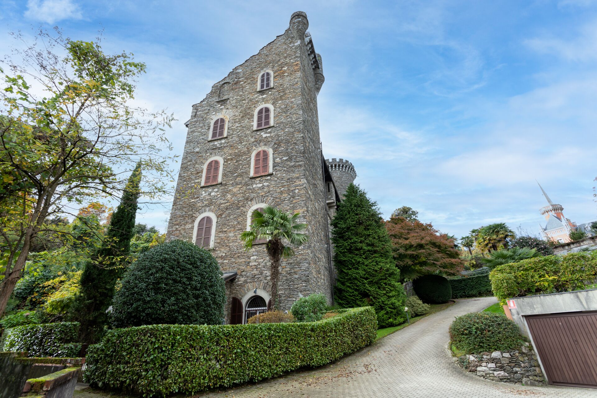 Ein Steinturm mit Bogenfenstern, umgeben von gepflegten Hecken und Bäumen.