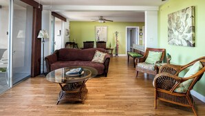 A spacious living room with a wooden floor, a glass coffee table, a brown sofa, and wicker chairs.