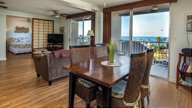 A spacious living room with a wooden dining table, wicker chairs, a sofa, and a television. There is a balcony with a view of the ocean.