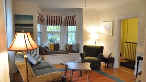 Uma sala de estar acolhedora com um sofá, poltrona e uma mesa de centro de madeira. Há uma janela com cortinas listradas e um quadro na parede.