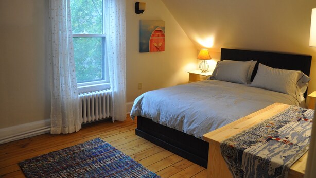 Um quarto com cama, mesa de cabeceira, luminária, janela com cortinas, radiador e piso de madeira.
