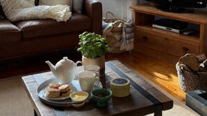Una sala de estar acogedora con un sofá de cuero marrón, una mesa de centro con una tetera, tazas y galletas, un televisor sobre un mueble de madera, y una planta grande.