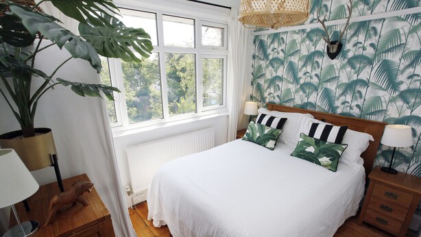 Un dormitorio con un ventanal grande, una cama con ropa de cama blanca, una mesita de noche de madera, una lámpara, una planta en maceta y una pared con diseño de hojas tropicales.