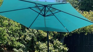 Un patio exterior con una mesa redonda y sillas de madera, rodeado de macetas con plantas y un paraguas grande.