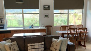 A living room with a brown sofa, wooden dining table, and chairs. There are two windows with blinds, a kitchen area with a sink, and a hanging light fixture. - Pyrenees Farm Stay Amphitheatre. (Pet friendly) Bed and Breakfast. (Amphitheatre)
