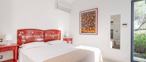 A bedroom with a large bed, a red nightstand, a white lamp, a wall-mounted air conditioner, a colorful abstract painting, and a large window with a view of greenery.