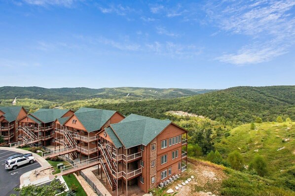 A multi-story hotel with a brick exterior and green roofs, surrounded by lush greenery and hills.