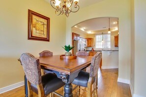 A dining room with a wooden table and chairs, a chandelier, and a kitchen in the background.
