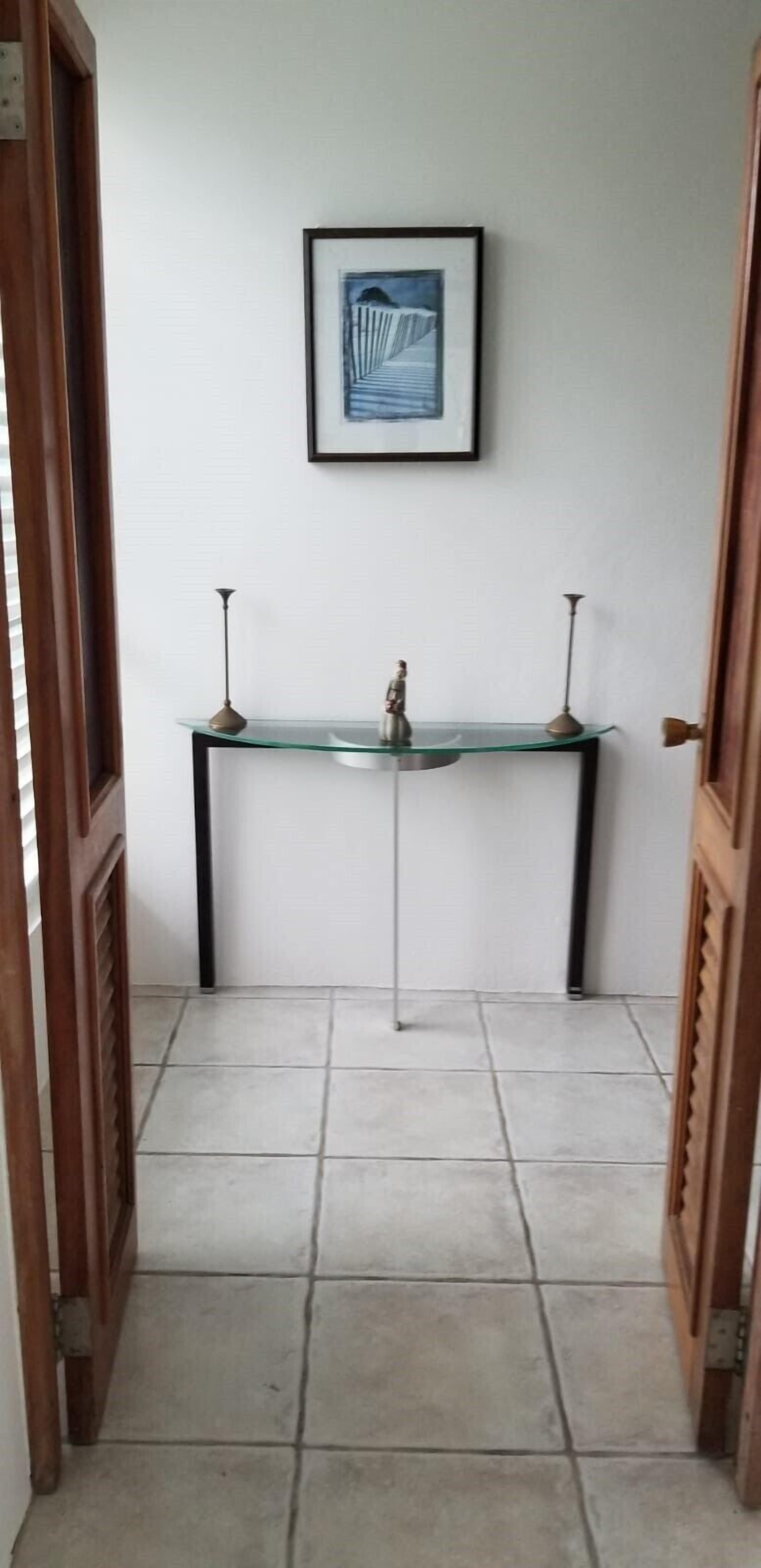 Un pasillo con una mesa consola de vidrio, una obra enmarcada encima y suelo de baldosas.