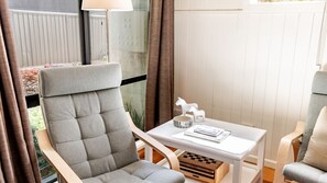 Une pièce moderne avec un fauteuil à revêtement gris, une petite table blanche agrémentée d’objets décoratifs, et une vue sur une clôture et la verdure à l’extérieur.