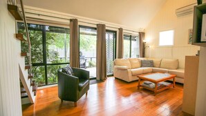 A spacious living room with a wooden floor, a sofa, a coffee table, and a chair. Large windows with curtains allow natural light and views of the outdoors. - ALLAMBIE COTTAGES - VILLA 3: Bright (Bright)
