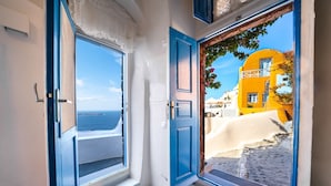 A room with two open windows, one with a view of the sea and the other with a view of a yellow building.