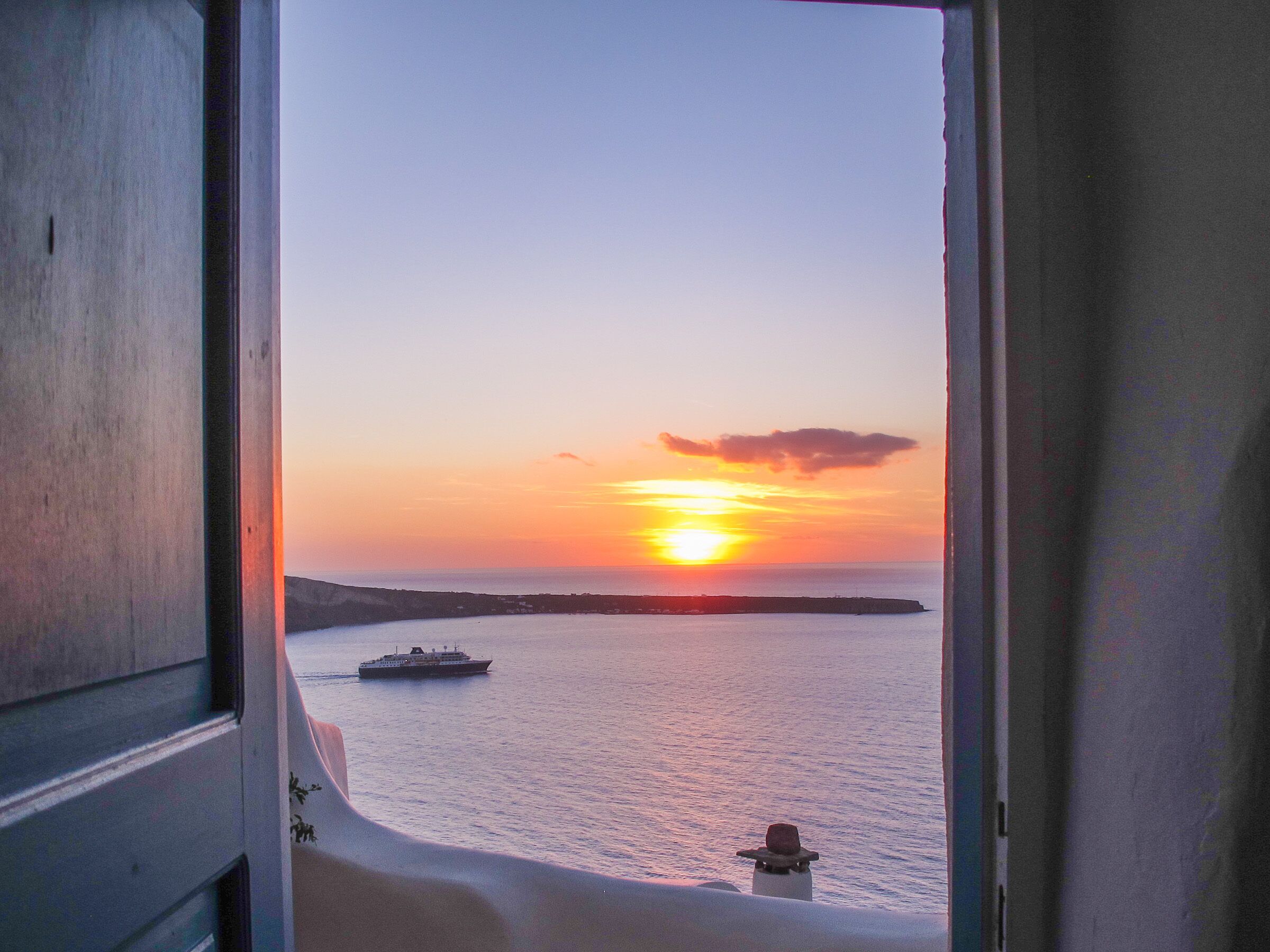 A sunset view from a window overlooking a body of water with a ship and a distant shoreline.