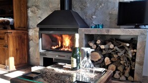 Ein Kamin aus Stein mit einem modernen Holzofen, ein Glastisch mit Flasche und Gläsern sowie ein Fernseher über dem Kamin.