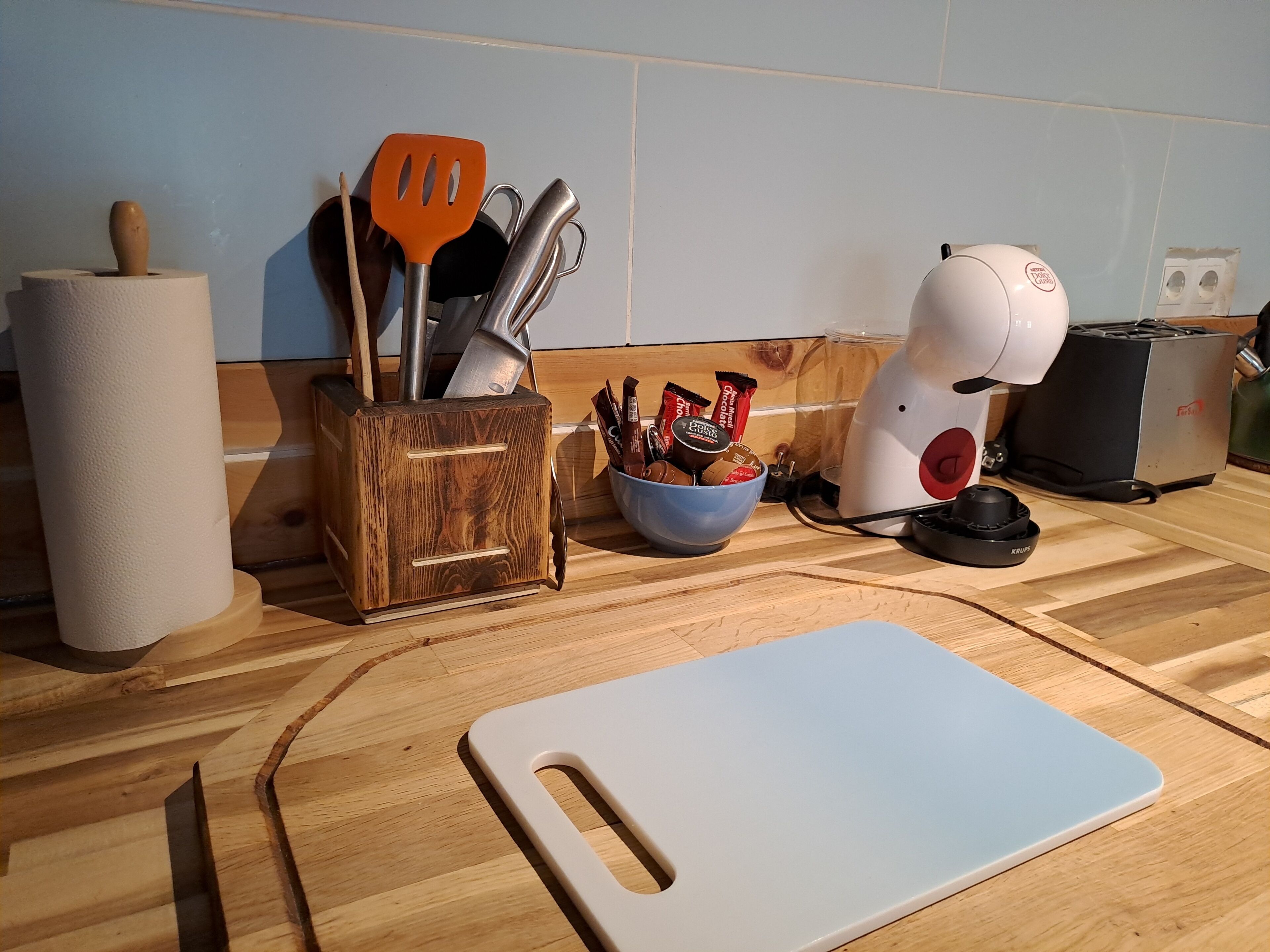 Una encimera de cocina con una tabla de cortar, un estante de madera para especias, un tazón de barras de chocolate y una cafetera.