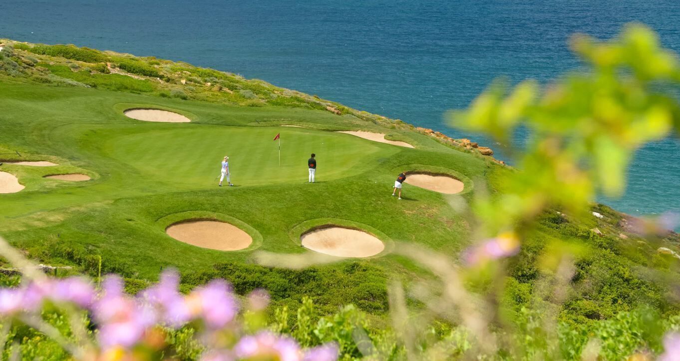 A golf course with bunkers and a flag, overlooking a body of water.