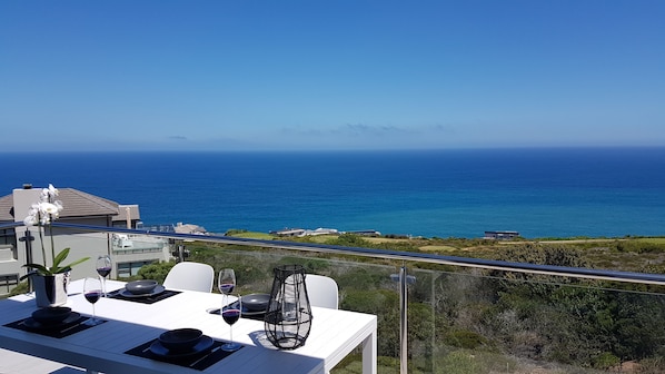 A balcony with a table set for dining, overlooking a coastal landscape with the ocean and distant buildings.