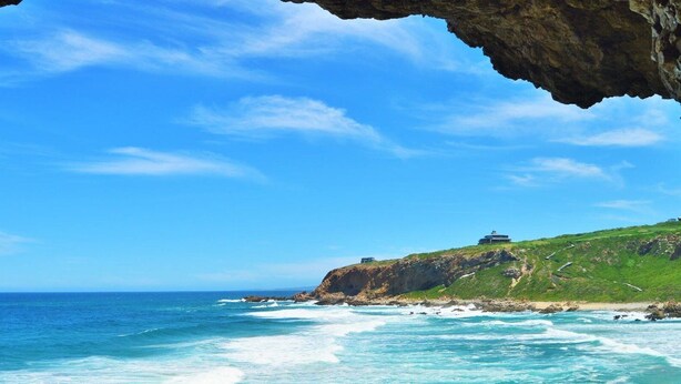 A coastal landscape with a building on a grassy hill overlooking the ocean.