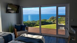 A bedroom with two beds, a TV mounted on the wall, and a large sliding glass door leading to a balcony with a view of the ocean and greenery.
