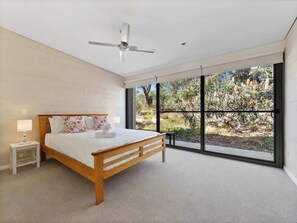 Un dormitorio con cama, mesita de noche, ventilador de techo y ventanas amplias.