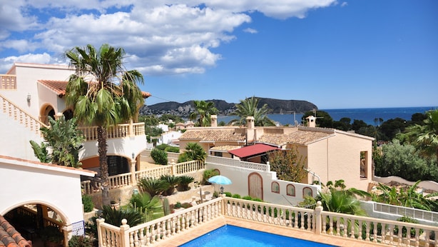 Ein Poolbereich mit Blick auf das Meer, umgeben von Gebäuden im mediterranen Stil, darunter eine Palme und ein Gebäude mit rotem Dach.