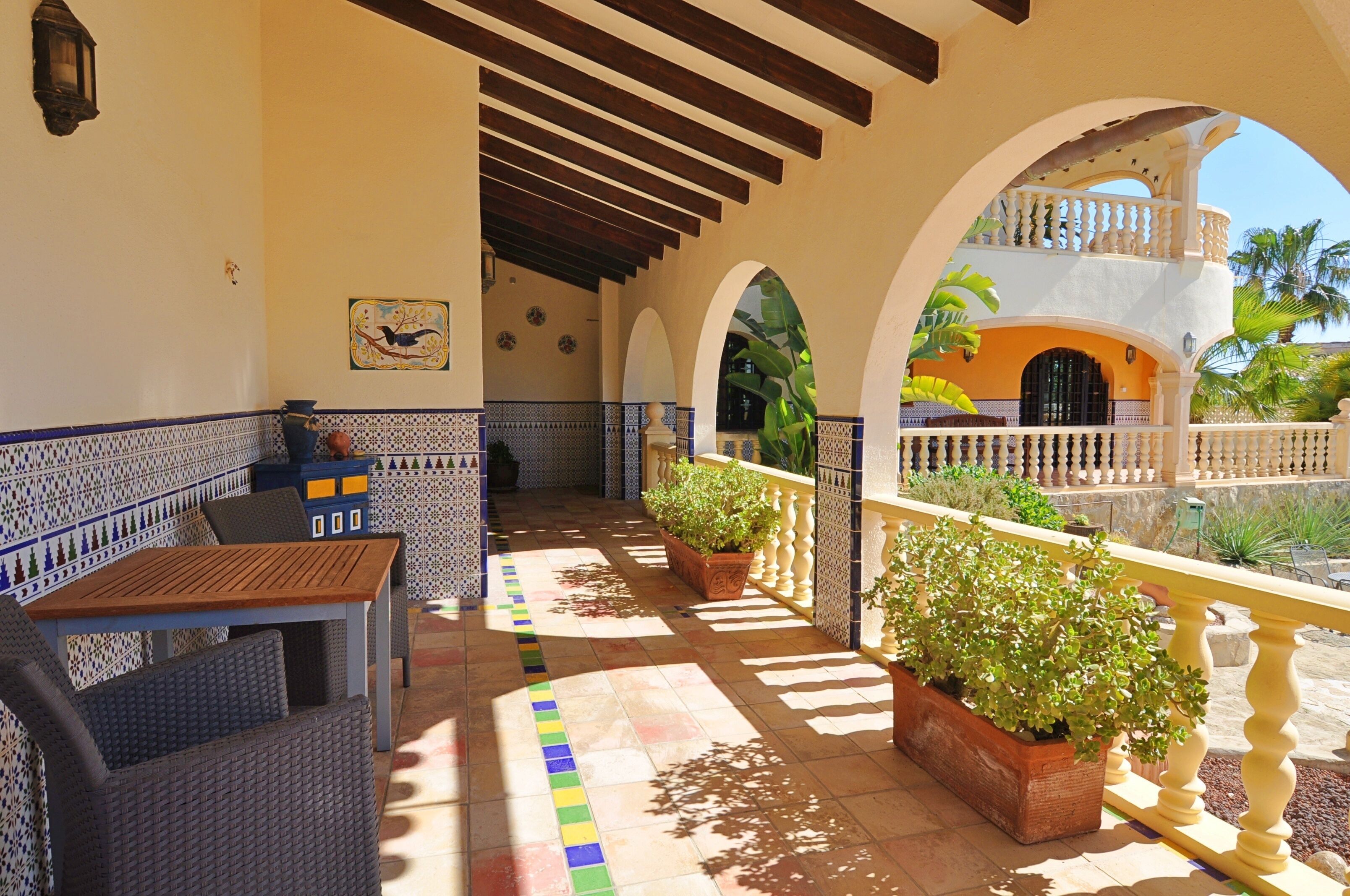 A covered patio with a wooden table and wicker chairs, potted plants, and a view of a building with a balcony.