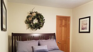 Ein ordentlich bezogenes Bett mit einem gestreiften Bettbezug und zwei Kissen, ein hölzernes Kopfstück, eine Holztür, ein gerahmtes Bild an der Wand und ein dekorativer Kranz über dem Bett.