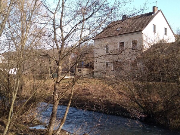 Un bâtiment blanc avec un toit recouvert de tuiles, entouré d’arbres sans feuilles et d’une rivière.
