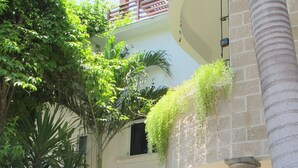 A two-story building with a brick facade, a wooden fence, and lush greenery including a palm tree and various plants. - Charming Penthouse, FREE INTERNET, CABLE and SAFE DEPOSIT BOX (Playa del Carmen)