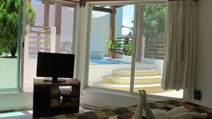 A room with a television on a wooden stand, a window with a view of a swimming pool and greenery, and a quilted bedspread. - Charming Penthouse, FREE INTERNET, CABLE and SAFE DEPOSIT BOX (Playa del Carmen)