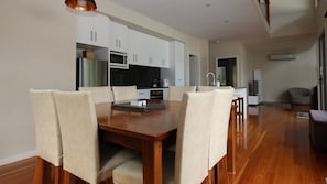 A modern dining area with a wooden table and six chairs, a kitchen with white cabinets, and a living area with a sofa and a TV.
