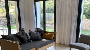 Ein modernes Wohnzimmer mit einer Couch und Sesseln, großen Fenstern mit Vorhängen und Blick ins Freie.
