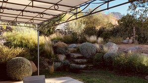 Eine Gartenpatio mit Holztisch und -stühlen, einer Metall-Pergola und einem angelegten Garten mit Steinen und Sträuchern.