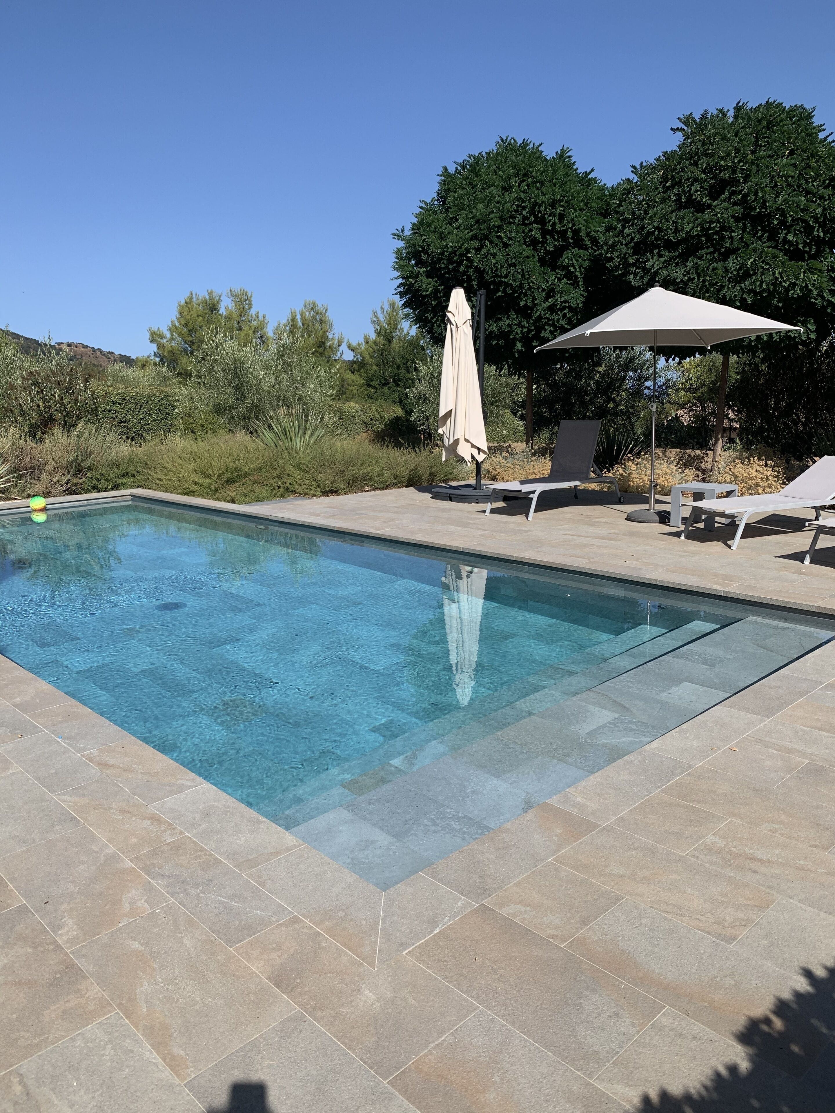 A rectangular swimming pool with clear blue water, surrounded by beige tiles. There are two lounge chairs and a single umbrella.