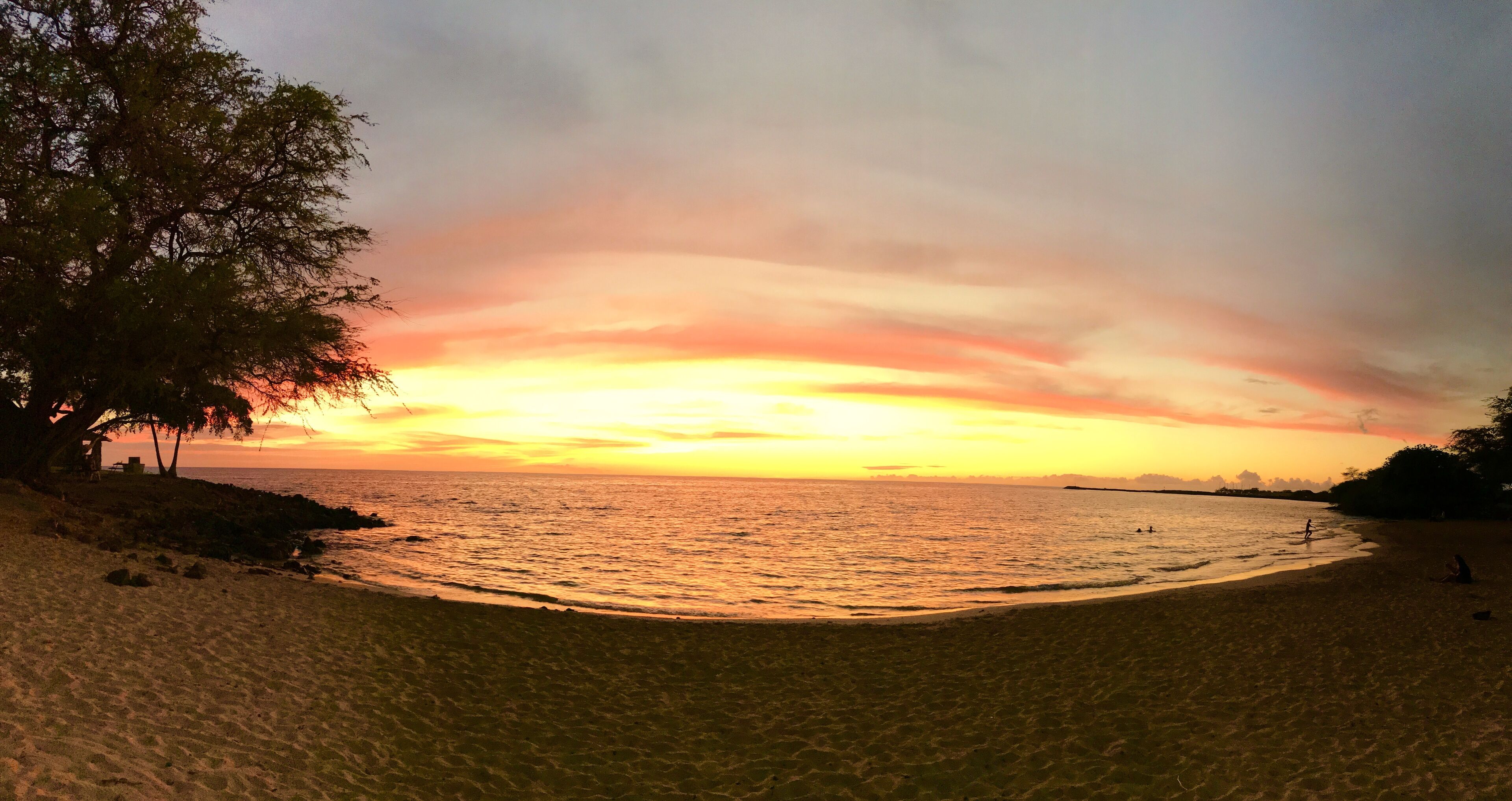 Una playa con un atardecer, mar tranquilo y algunas personas en la orilla.