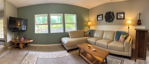 A living room with a beige sofa, a wooden coffee table, a TV mounted on the wall, and a window with a view of greenery.