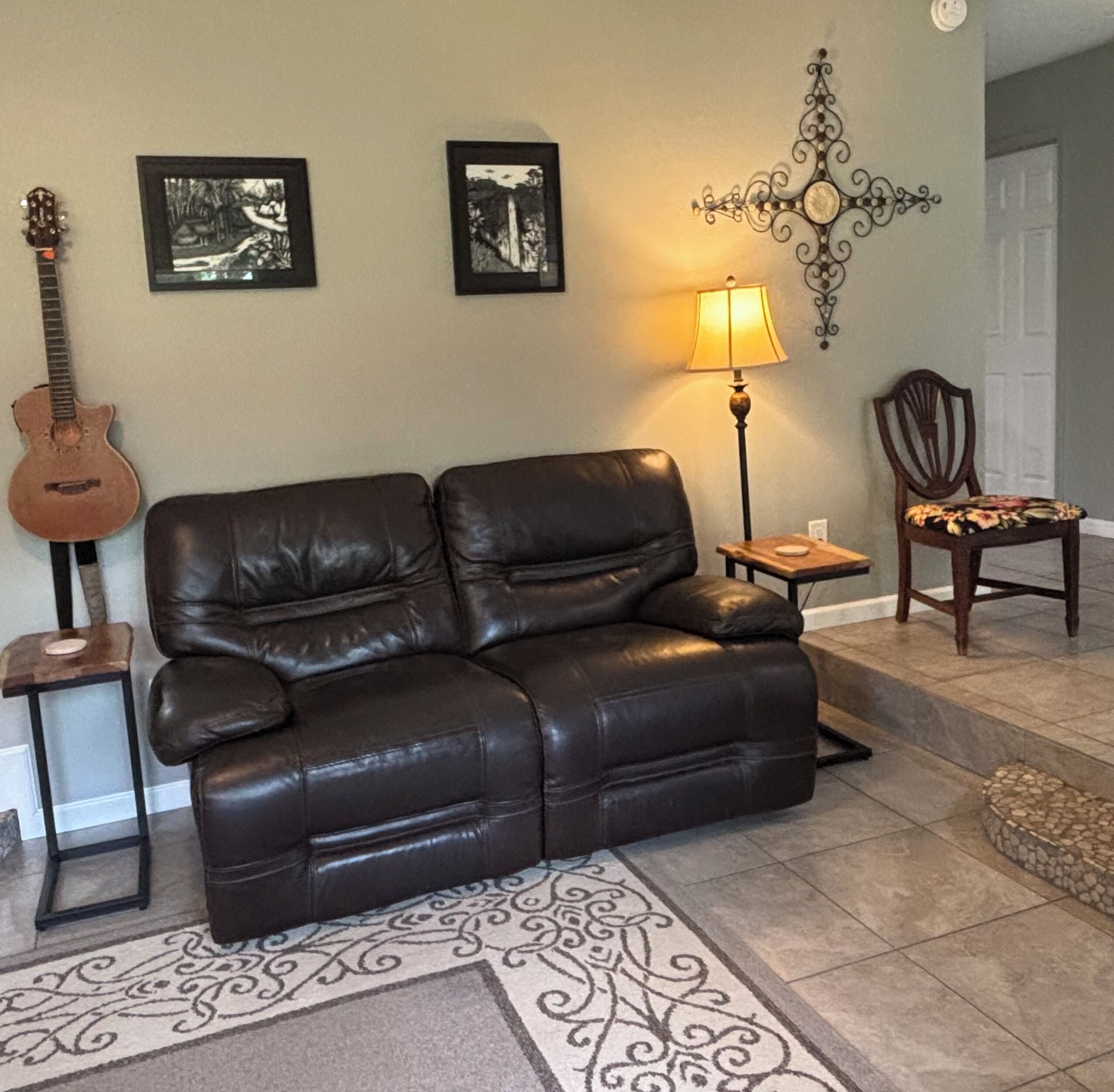 Un salon avec un canapé en cuir, une guitare, deux tableaux encadrés, une lampe, un fauteuil et un tapis à motifs.