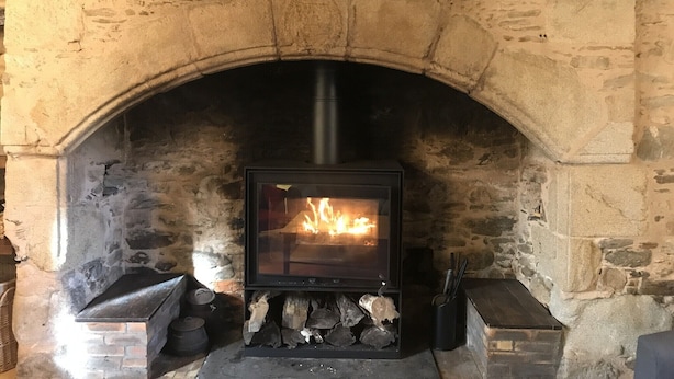 Une cheminée en pierre avec un poêle à bois moderne, du bois de chauffage rangé en dessous et une cheminée au-dessus.