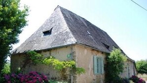 Une maison de campagne au toit à forte pente, entourée d’une végétation luxuriante et de plantes en fleurs.