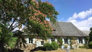 Une maison en pierre avec un toit en pente, des volets blancs et un jardin agrémenté d’un grand arbre en fleurs.