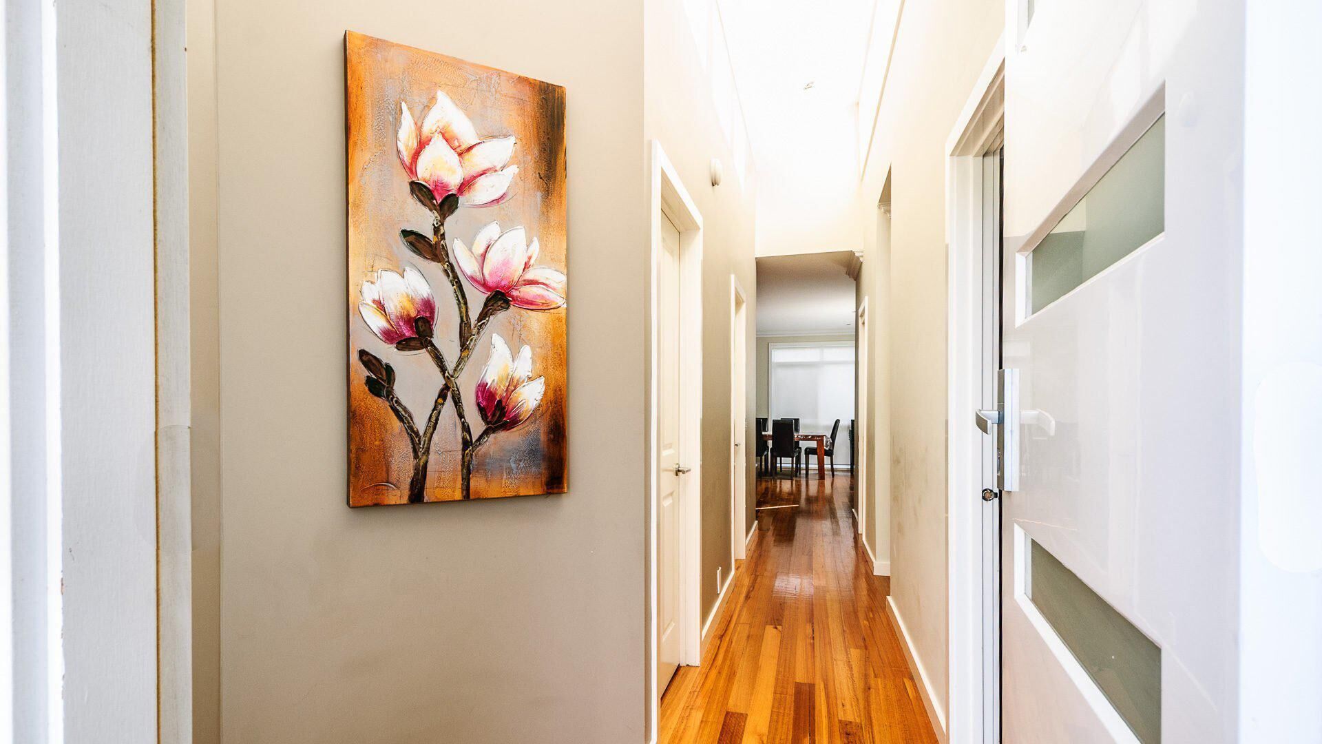 A hallway with wooden flooring, a painting of magnolia flowers on the wall, and a dining area in the distance.