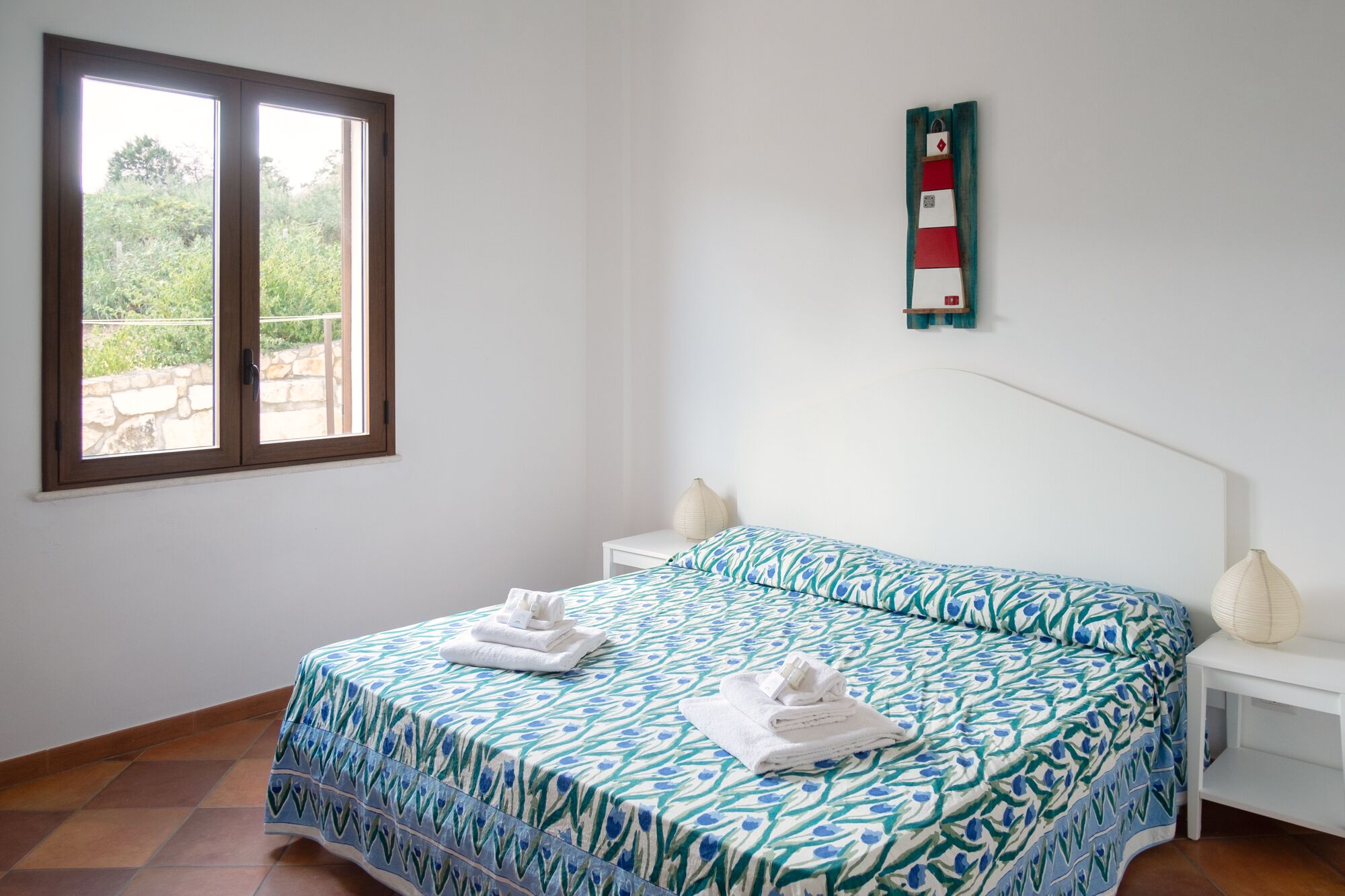 A bedroom with a bed, a nightstand, a lamp, a window, and a wall-mounted decoration.