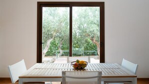 Un coin repas avec une table et des chaises, un panier de fruits sur la table et une vue sur les arbres à travers une porte vitrée.
