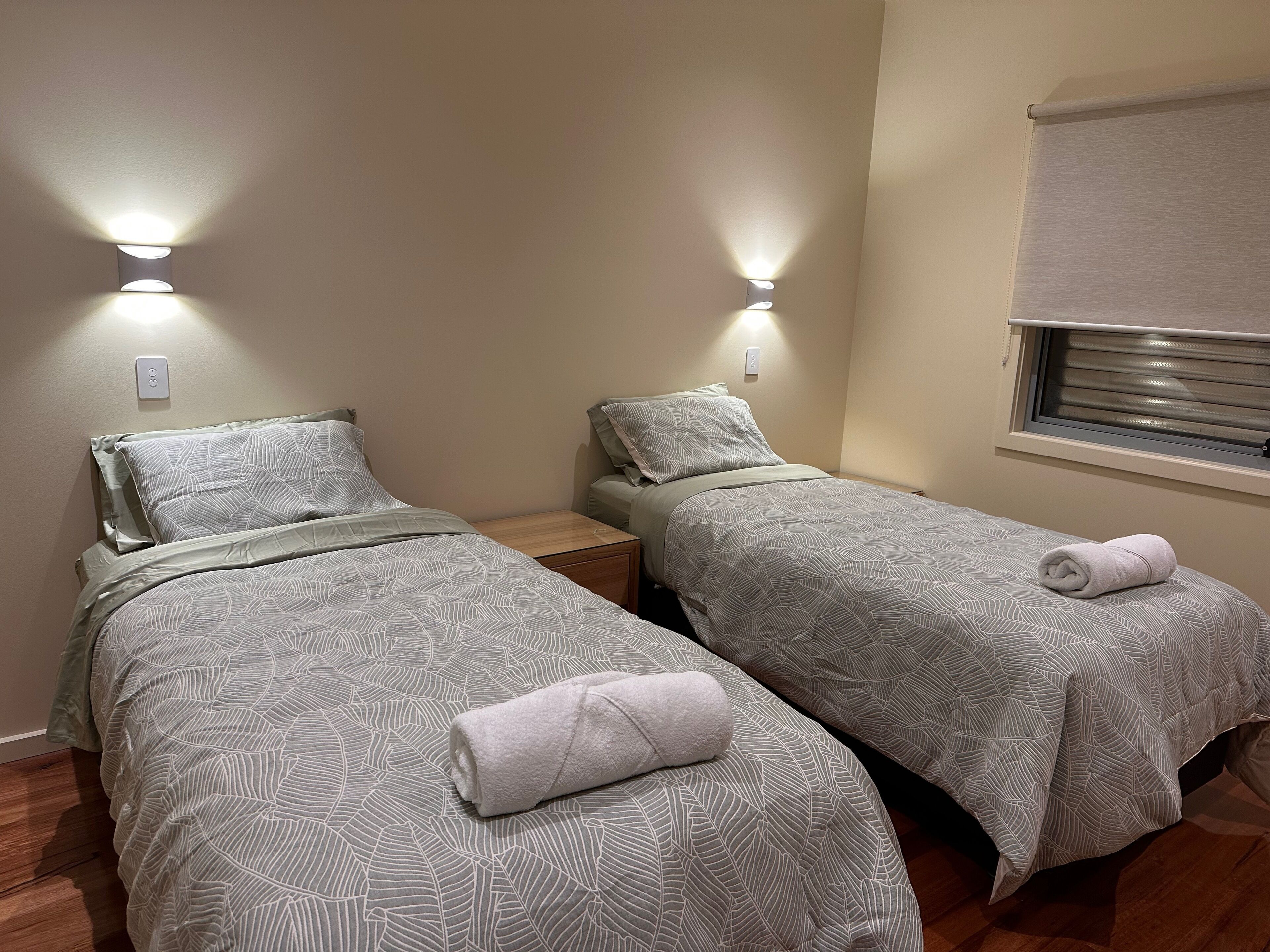 A twin room with two beds, each with patterned bedding and a rolled towel. There are wall-mounted lights and a window with blinds.
